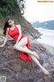 A woman in a red dress sitting on a rock by the ocean.